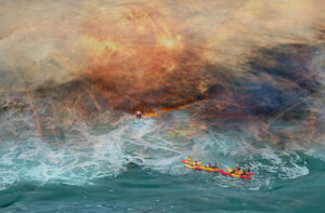 People in kayaks in rough seas try to rescue a boy caught on a branch. The background is turbulent.